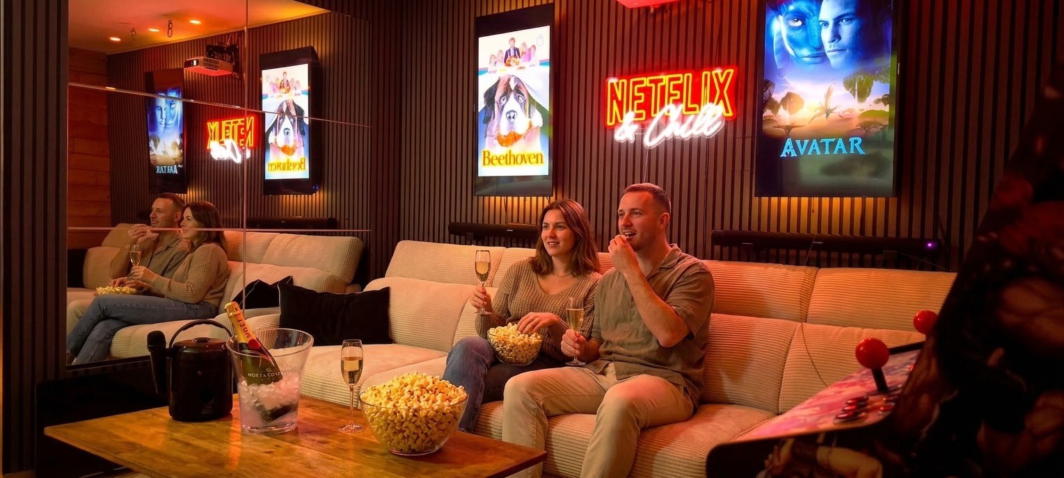 Un couple sur le grand canapé de la ciné room, coupes de champagne et popcorn, sous le néon Netflix & Chill et les affiches lumineuses