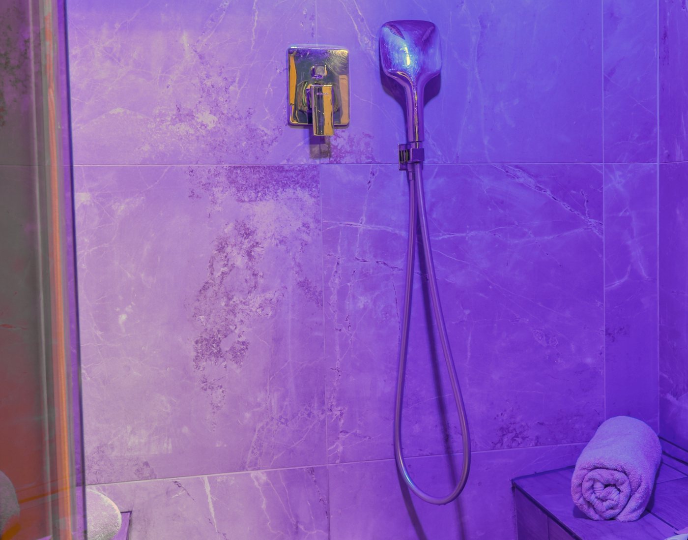 Le hammam éclairé en violet : murs en marbre, douchette chromée, banc en bois avec serviettes roulées et produits d'accueil