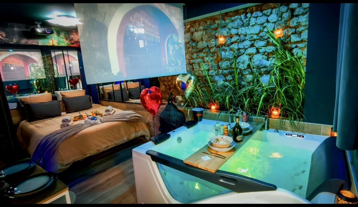 Le jacuzzi éclairé avec son plateau de bain dressé, à côté du lit, sous l'écran géant et devant le mur en pierre