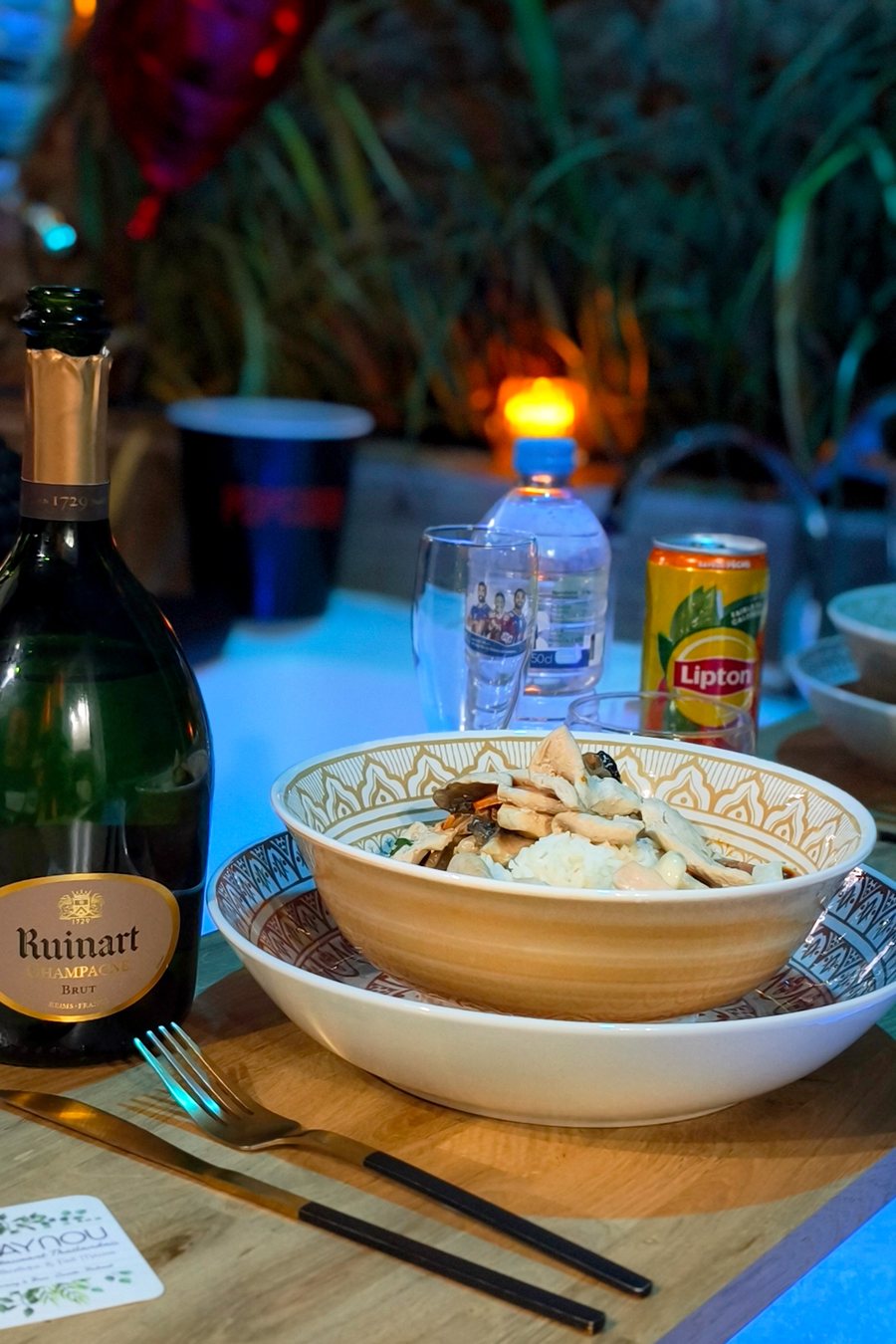 Le dîner servi sur le plateau en bois du jacuzzi : bol de riz au poulet, champagne, verres, bougie et ballon cœur devant le mur en pierre
